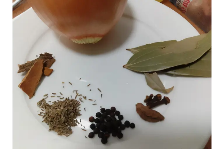 Whole spices and onion used in chicken pulao recipe including bay leaves, cinnamon, cumin seeds, black pepper, and cloves.